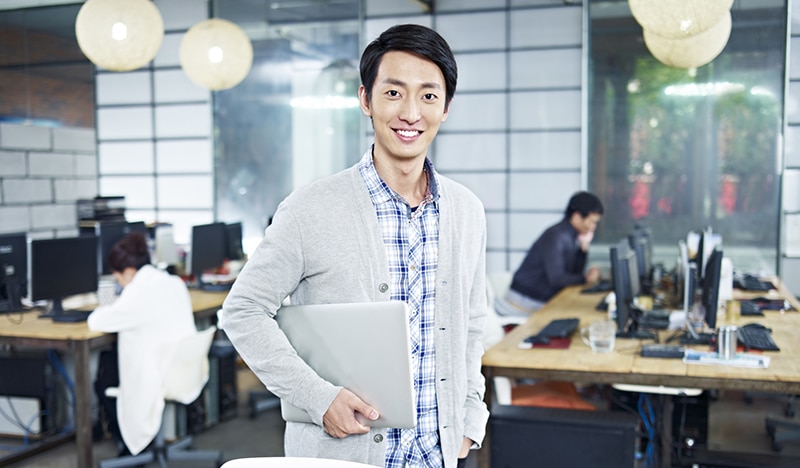 Confident young asian entrepreneur standing in own company with laptop computer looking at camera smiling.