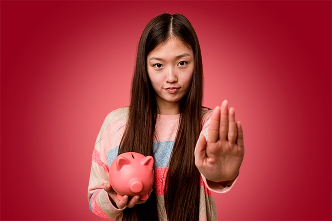 Young asian woman holding a piggy bank standing with outstretched hand showing stop sign, preventing you.