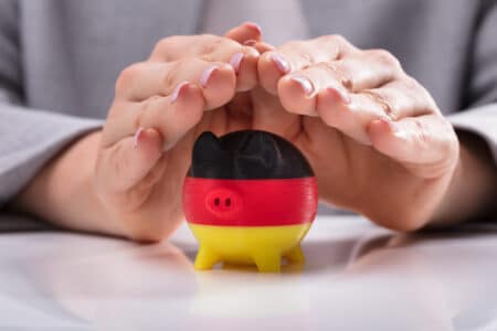 Close-up Of A Woman's Hand Protecting Piggybank Painted With German Flag On Desk
